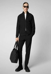 Giacca nera con zip, t-shirt bianca, pantaloni neri e stivali neri. Tiene una borsa nera con doppi manici. Texture liscia, design minimalista.