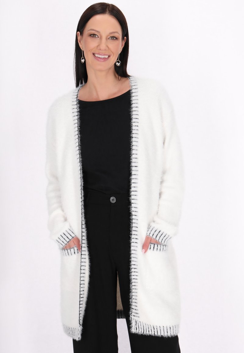 Strickjacke in Weiß mit schwarzem Streifenrand, offenen Front, langen Ärmeln und zwei Vorderaschen. Weiche, flauschige Textur.