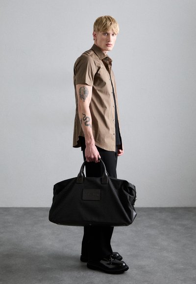 Sac de sport noir avec accents en cuir, tissu texturé et un patch à l'avant. Le modèle porte une chemise marron à manches courtes et un pantalon noir.