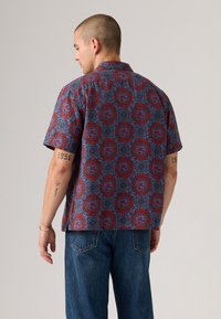 Man with short hair wearing a red and blue patterned short-sleeve shirt and blue jeans, viewed from the back against a plain background.
