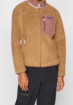 Veste en polaire beige avec poche poitrine mauve et fermeture éclair, portée avec un pantalon noir doté de poches avant zippées.