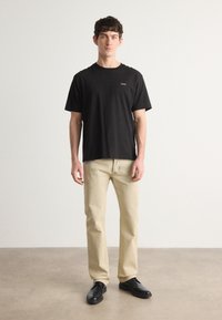 Tricou din bumbac negru cu guler rotund și un mic logo "BOSS", asortat cu blugi bej deschis și pantofi negri, stând pe un fundal neutru.
