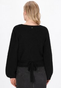 Frau mit blonden Haaren, die einen schwarzen, flauschigen Pullover mit einer Schleife am Rücken trägt, kombiniert mit dunkelgrauen Jeans, von hinten gezeigt.