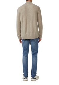 Mann in beigem Strickpullover, blauen Jeans und weißen Turnschuhen, der mit dem Rücken zu einer weißen Hintergrund steht.