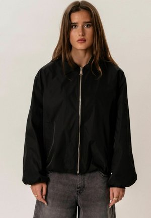 Fiatal nő hosszú barna hajjal, fekete cipzáras dzsekit és szürke farmert visel, egyszerű világos háttér előtt áll.