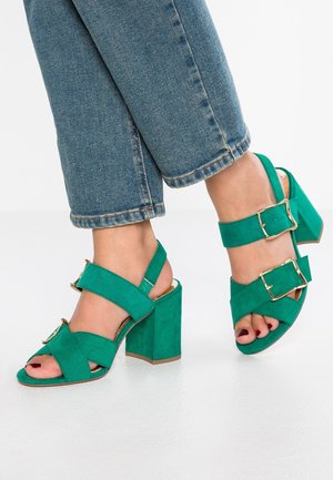 Vihreät mokkanahkaiset korkokengät, joissa on lohkarekorko, ristiriipusnauhat ja kultainen solki. Avoin varvasmuotoilu.