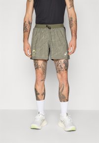 Grønnmønstrede shorts med elastisk liv og snor, med et logo og fjellgrafikk, iført hvite joggesko og sokker.