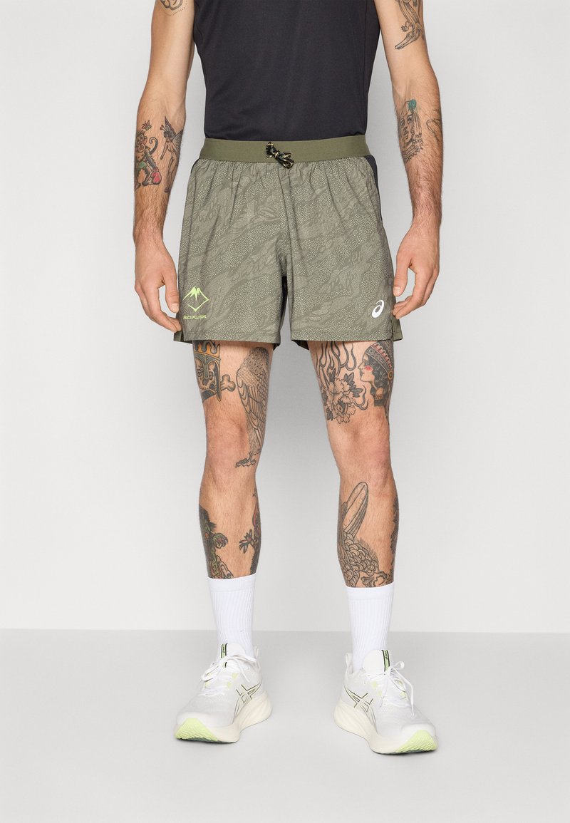 Grønnmønstrede shorts med elastisk liv og snor, med et logo og fjellgrafikk, iført hvite joggesko og sokker.