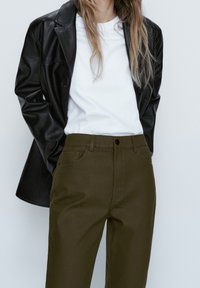 Schwarze Jacke aus Kunstleder, weißes T-Shirt und olivgrüne Hose. Die Jacke hat ein tailliertes Design; die Hose verfügt über Fronttaschen und einen Knopfverschluss.