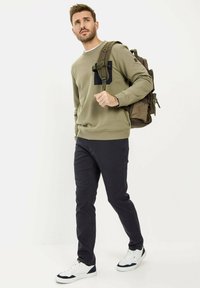 Olijfgroen sweatshirt met een zwarte zak, gecombineerd met donkere jeans en witte sneakers, aangevuld met een bruine rugzak.