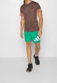 Camiseta deportiva marrón de manga corta, pantalones cortos verdes con un logo blanco y zapatillas deportivas negras con suela texturizada.