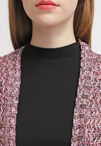 Detailní záběr dolní části obličeje a krku ženy, která má na sobě černý žebrovaný top a růžový, vícebarevný texturovaný pletený cardigan.