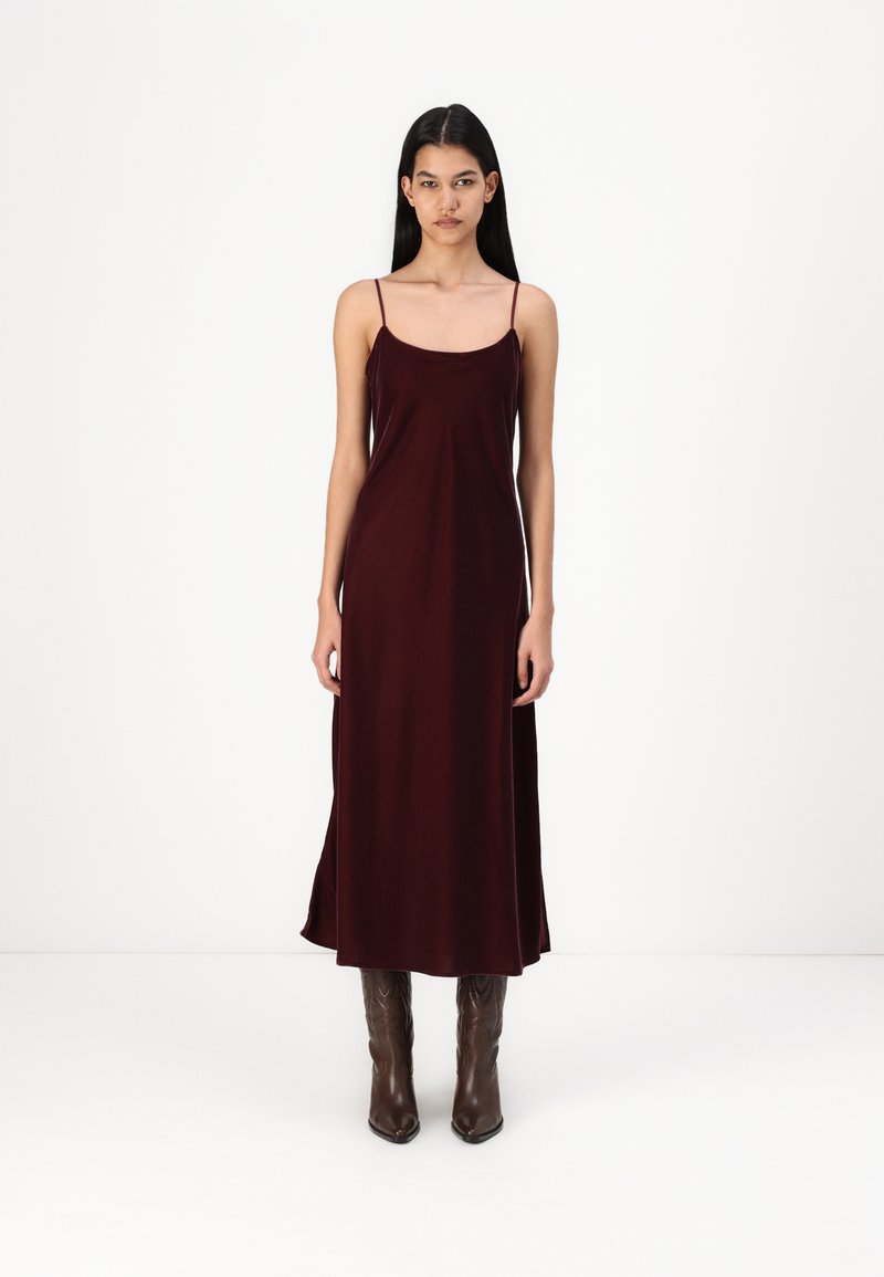 Rochie maxi fără mâneci, de culoare burgundă, cu un design fluid, textură fină și fante laterale. Asortată cu cizme maro, cu vârful ascuțit, până la genunchi.