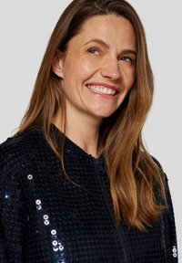 Schwarze, paillettenbesetzte Bomberjacke mit strukturiertem Material, verfügt über einen gerippten Kragen und einen Reißverschluss, getragen von einer Frau mit langen Haaren.