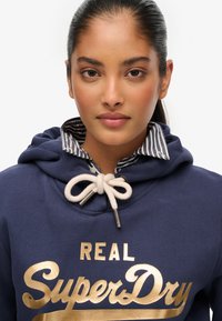 Sudadera azul marino con un estampado dorado de "SuperDry", con cierre de cordón y cuello interior a rayas. Material suave y corte relajado.