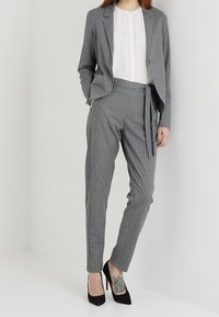 Donna che indossa un completo grigio a righe con pantaloni a vita alta, camicia bianca, tacchi alti neri e un tatuaggio ben visibile sulla caviglia, in piedi su uno sfondo bianco.