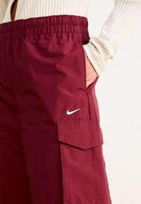 Burgunderfarbene Cargo-Hosen mit elastischem Bund, seitlicher Tasche und weißem Nike-Swoosh-Logo. Strukturierter Stoff mit sichtbaren feinen Linien.