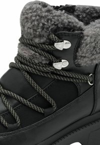 Schwarze Lederstiefel mit grauem, strukturiertem Fellfutter, ausgestattet mit schwarz-grauen, gemusterten Schnürsenkeln, silbernen Ösen und einer dicken Gummisohle.