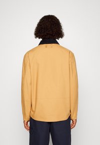 Yellow cotton jacket with a black collar, featuring a straight cut, long sleeves, and two looped accents at the back.