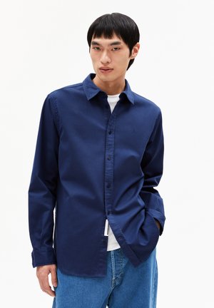 Navy blue long-sleeve button-up shirt made of smooth fabric, featuring a collar and a small embroidered logo on the chest. Worn with loose denim jeans.