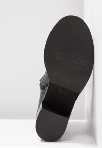 Semelle de chaussure en cuir noir avec un talon texturé et un devant lisse, positionnée debout contre des surfaces blanches, montrant le logo de la marque et la taille 37.
