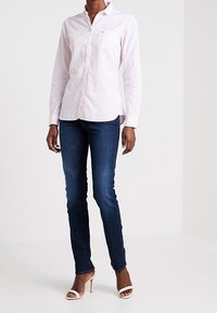 Camicia col colletto di un leggero rosa, con tasca frontale, abbinata a jeans slim blu scuro e tacchi bianchi con cinturini. Tessuto morbido, design aderente.