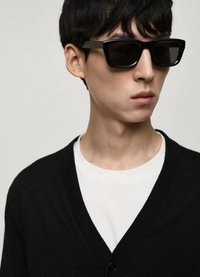 Svarta rektangulära solglasögon med mörka linser, bärs av en modell i en svart cardigan över en vit t-shirt. Slät textur och modern design.