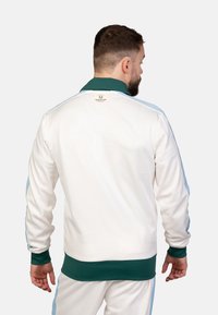 Weiße Sportjacke mit grünem Kragen und Bündchen, mit blauen Seitenstreifen und einem kleinen Markenlabel am hinteren Kragen.