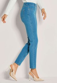 Frau in engen hellblauen Jeans und weißen spitzen High Heels mit transparenten und Perlen-Details vor einem schlichten hellen Hintergrund.