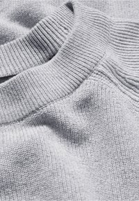 Gros plan sur un tissu tricoté doux et gris clair montrant un col côtelé et une texture de tricot détaillée avec des plis et des ombres délicats.
