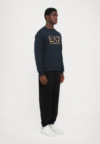 Navy sweatshirt with gold logo, paired with black joggers and white sneakers. Lightweight fabric, long sleeves, ribbed cuffs and hem.