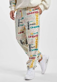 Multi-colored track pants featuring a cream background with repeated "DNGRS" text in various fonts. Worn with white sneakers.