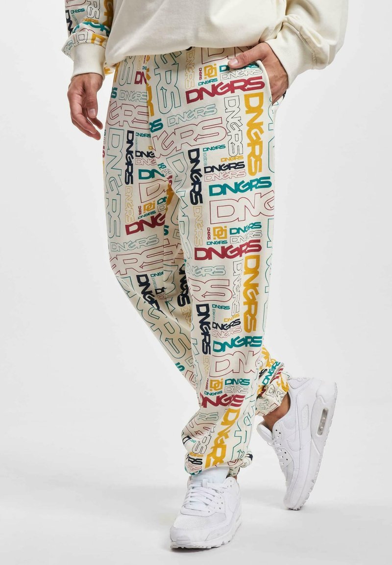 Multi-colored track pants featuring a cream background with repeated "DNGRS" text in various fonts. Worn with white sneakers.
