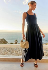 Frau in einem ärmellosen, navyblauen Maxikleid, die eine geflochtene Handtasche hält und auf einer Terrasse steht, mit einem felsigen Strand und dem Ozean im Hintergrund.