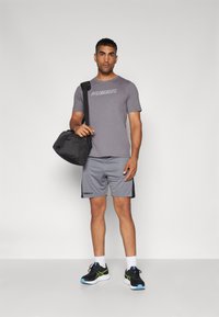 Camiseta atlética gris con el logo "HUMMEL", pantalones cortos grises con acentos laterales, zapatillas deportivas negras y una bolsa de deporte negra.
