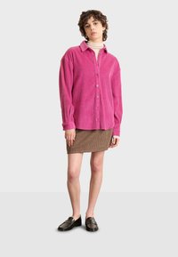 Camisa de pana rosa con botones en el frente, mangas largas y puños abotonados, combinada con una falda marrón texturizada y mocasines negros.