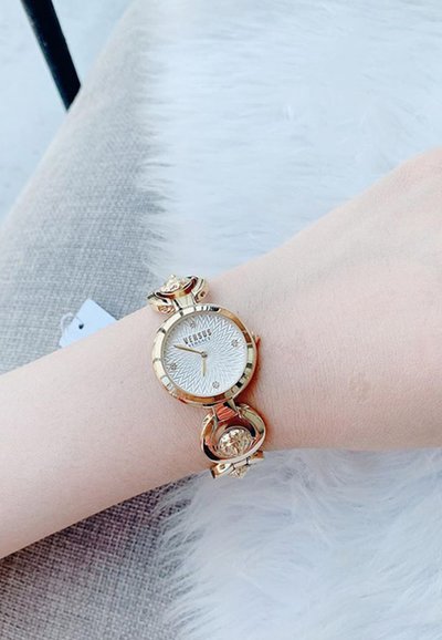Reloj en tono dorado con una esfera blanca texturizada, pulsera decorativa de eslabones de cadena y acentos florales en los eslabones. Muestra la hora con precisión.
