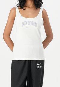 Bílý bavlněný tílko s širokými ramínky, s logem "NIKE SPORTS" v purpurové barvě a symbolem Nike swoosh. Kombinováno s černými sportovními šortkami.