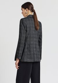 Donna con una lunga coda di cavallo marrone, che indossa orecchini a cerchio argentati, un blazer grigio a scacchi e pantaloni neri a gamba larga, voltata di spalle.