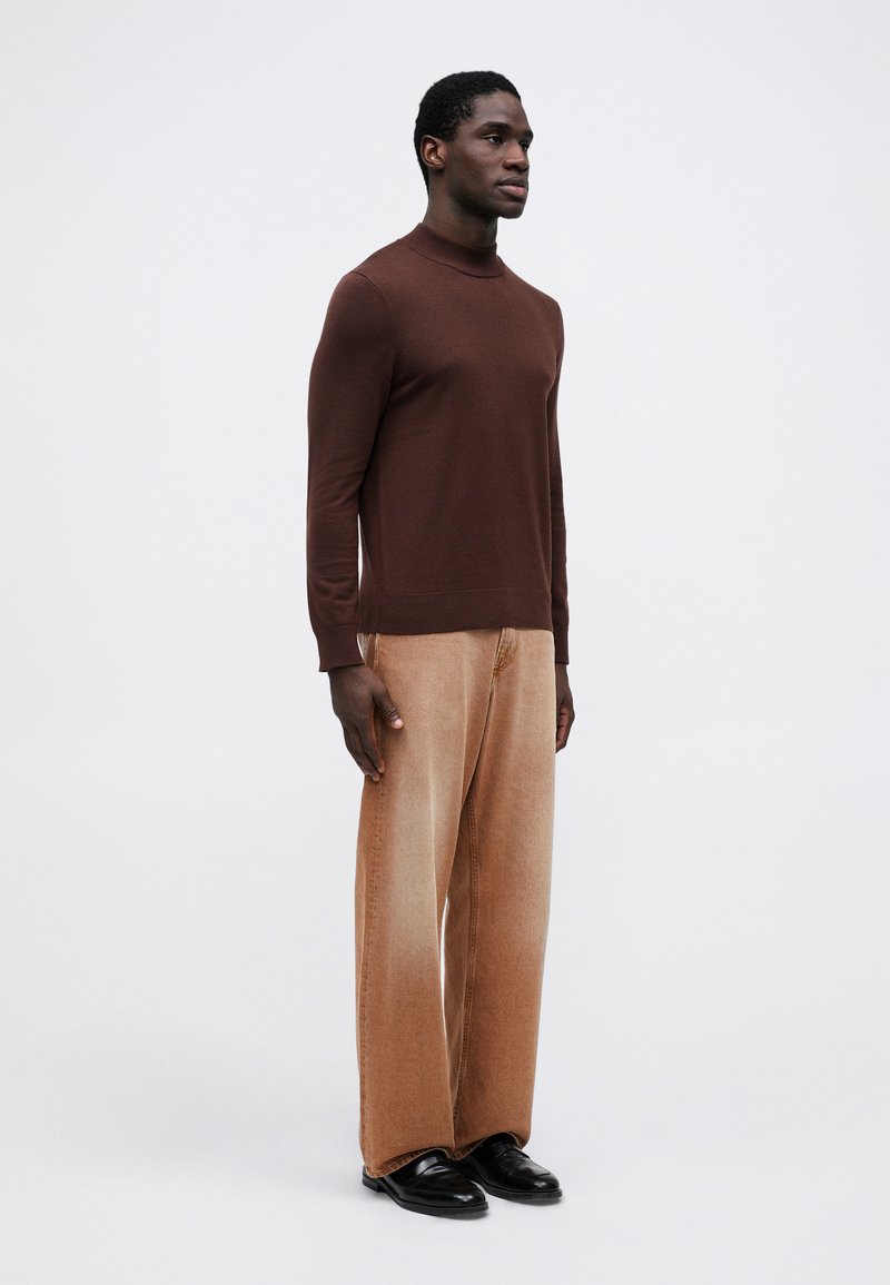 Brun striktrøje med høj hals, parret med lysebrune vidde bukser og sorte sko. Modellen står i profil.