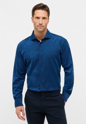 Blauwe, lange mouwen shirt van een zachte stof met een knoopsluiting aan de voorkant, puntige kraag en contrasterende knopen; gecombineerd met donkere broeken.