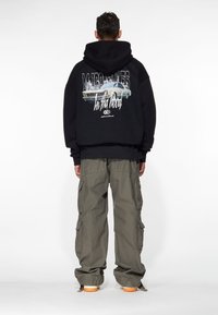 Zwarte hoodie met een afbeelding van een klassieke auto op de achterkant, gecombineerd met olijfgroene cargobroek met meerdere zakken. Tekst op de hoodie: "In tha Wood."