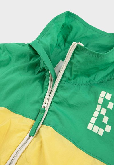 Veste verte et jaune avec une fermeture éclair à l'avant et un motif carré pixélisé blanc sur la partie supérieure verte.