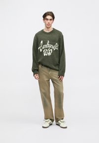 Pulover verde tricotat, cu text alb pe partea din față, asociat cu pantaloni bej cu croială largă și adidași crem cu accente negre.