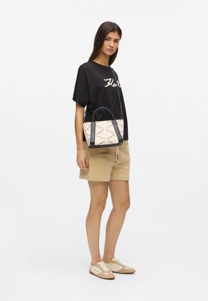Woman stands wearing black logo t-shirt, beige shorts, beige sneakers, and crossbody beige and black handbag with printed text pattern.