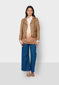 Braune Lederjacke mit Reißverschlüssen, Creme-zu-Braun Ombre-Oberteil, breite blaue Jeanshose und beige Rutschsandalen. Modell steht aufrecht.