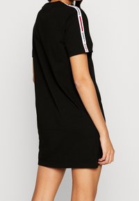 Svart T-shirt klänning med avslappnad passform, korta ärmar och kontrasterande vita och röda märkesränder på axlarna. Slät bomullstextur.