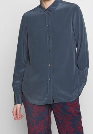 Personne portant une chemise bleue foncée à manches longues boutonnée avec un pantalon à motifs rouge foncé et bleu, debout, une main détendue le long du corps.