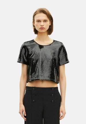 Jongere in een zwart, glinsterend crop top en hoge zwarte broek met dubbele knoopdetails, staande tegen een effen achtergrond.