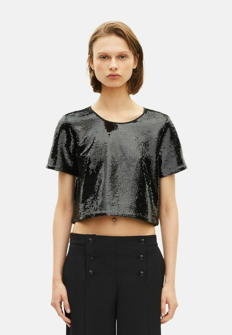 Giovane in top corto nero con pailette e pantaloni neri a vita alta con dettagli a doppio bottone, in piedi su uno sfondo neutro.